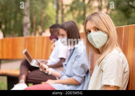 Young blond woman in protective mask looking at you while sitting on bench against two millennials using smartphone and laptop Stock Photo