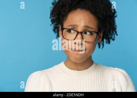 Close up studio portrait of shy awkward young Afro American woman wear glasses white sweater, biting lips feeling embarrassed, confused and nervous, l Stock Photo