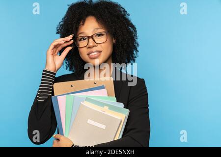 Happy teacher or businesswoman in blazer, wear glasses, holding notebooks, documents and folder, smiling looking at camera isolated on blue studio wal Stock Photo