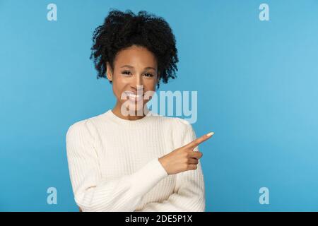 Smiling dark skinned woman with curly hair wear white jumper pointing with finger, showing blank copy space for advertising, offering, product, promot Stock Photo