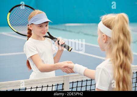 Happy teenage girl in activewear holding tennis racket while shaking hand of her friend over net against field before new training on stadium Stock Photo