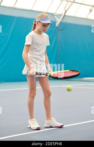 Youthful blond girl in white activewear holding tennis racket while standing on stadium and pushing ball during training in front of camera Stock Photo
