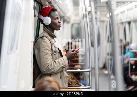 Afro-American passenger man in red hat, trench coat stand in subway train, using mobile smart phone, listens to music with wireless headphones in publ Stock Photo