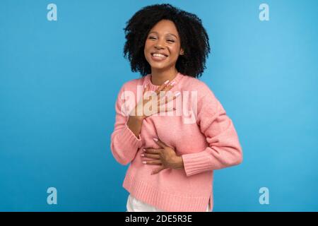 Laughing mixed race millennial woman with curly hair in casual pink jumper, keeps hand on chest, having fun, isolated on studio blue background. Overj Stock Photo