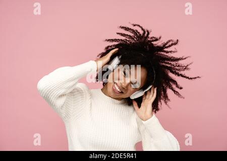Energized biracial woman sing along, smile broadly, wears wireless headphones, has good mood, isolated on pink background. Happy dark skinned curly gi Stock Photo
