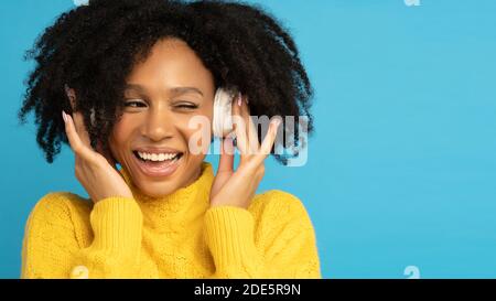 Smiling mixed race woman in yellow jumper touching wireless headphones, listening to music, has good mood, winks and looks at blank space for advertis Stock Photo