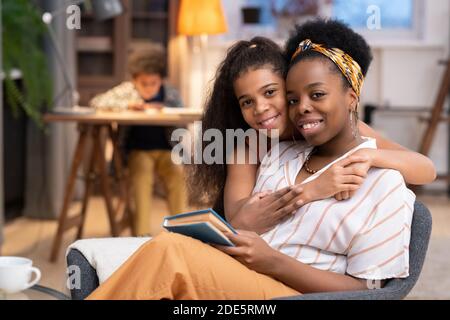Happy African girl with toothy smile embracing her mother sitting in armchair in front of camera on background of cute boy doing homework Stock Photo