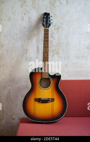 Acoustic vintage black and orange guitar on red couch at loft style apartment over concrete wall . Vertical. Stock Photo