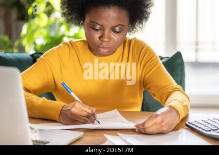 Black woman employee or manager remote working at home during lockdown, makes notes with pen, works with paper documents. Office worker during a pande Stock Photo