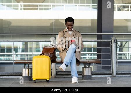 Smiling Afro-American traveler man with yellow suitcase sitting on bench in airport terminal or railway station, using mobile phone, calling taxi, wai Stock Photo