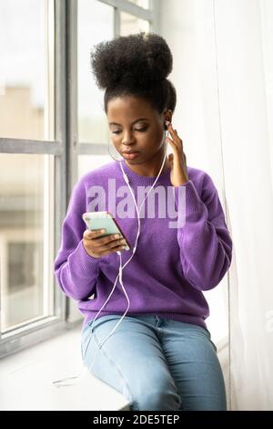 African American millennial young woman with afro hairstyle wear sweater, sitting on windowsill, listens to music with wired headphones on mobile smar Stock Photo