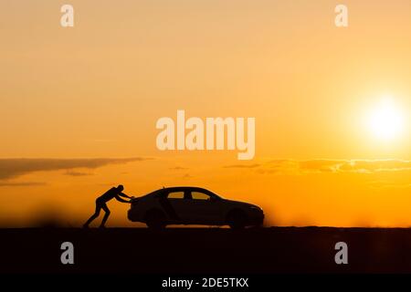 Transportation trouble. Silhouette of man driver pushing his car along on an empty road after breakdown at sunset, copy space, side view. Stock Photo