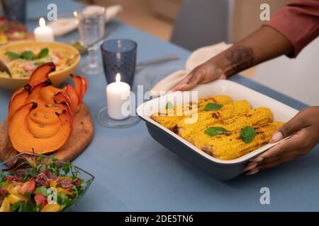 Hands of young African woman putting bowl with grilled corn on festive table next to burning candle, baked pumpkin and vegetable salad Stock Photo