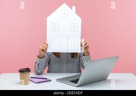 Woman in striped shirt hiding face behind paper house, working on laptop from home, notepad and paper cup lying on table, freelancer. Indoor studio sh Stock Photo