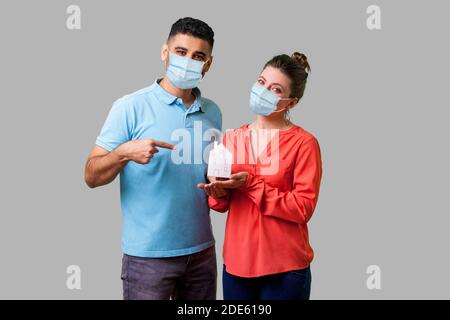 Portrait of happy couple with surgical medical mask looking at camera with happiness, man pointing at house in woman hands. stay home, stay safe on qu Stock Photo