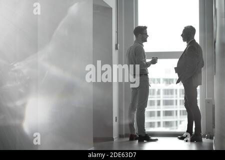 Black and white photo of two businessmen standing in the modern office against panoramic window and discussing something while having coffee break Stock Photo