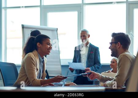 Two cheerful diverse office workers discussing something and smiling while having a meeting with colleagues in the modern office Stock Photo