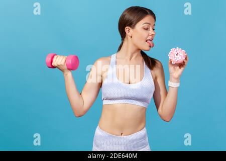 Athletic woman in white sports tights and top holding dumbbell and donut, showing tongue going to lick it, keeping diet going for sport. Indoor studio Stock Photo