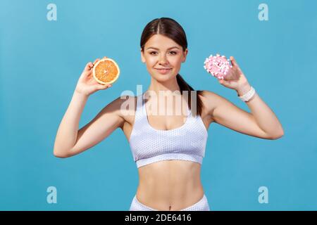 Healthy slim woman in sportswear holding orange and donut in hands, making choice between healthy and junk food nutrition. Indoor studio shot isolated Stock Photo