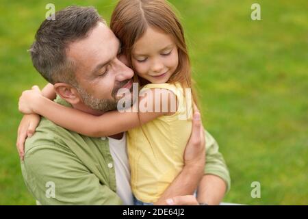 Portrait of happy young father and adorable little daughter hugging each other, standing with eyes closed while spending time in the green park on a Stock Photo
