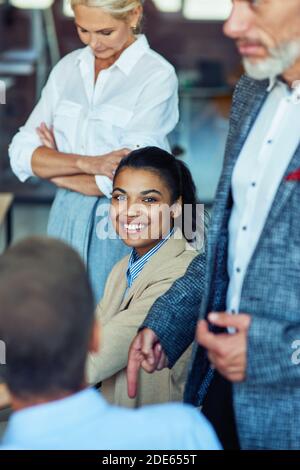 Young cheerful mixed race woman, female office worker smiling at camera while having a meeting with colleagues Stock Photo