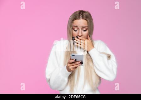 Anxious woman covers mouth in shock, holding and looking at mobile smart phone, surprise reaction on her face. Awkwardness gestures, expression concep Stock Photo