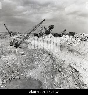 1950s, historical, mining, picture shows a large stone or rock quarry, with mechanical dragline excavator, with excavating grab bucket or shovel on a pulley system moving rocks at the side of the site. Quarrying is a multistage process involving the initial breaking up of the rock or earth and then its extraction from the ground. Stock Photo