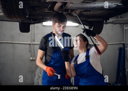 A female engineer and a workman in uniform are repairing car brakes. They use a flashlight Stock Photo