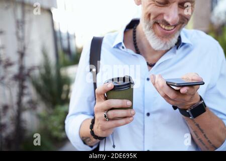 Cropped shot of modern middle aged businessman smiling while using his smartphone, walking outdoors with a cup of coffee Stock Photo
