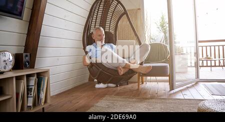 Happy middle aged caucasian businessman in casual clothes sitting on swing chair at his modern apartment, working online on laptop Stock Photo