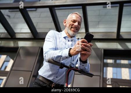 Generating ideas. Cheerful middle aged businessman smiling while using his smartphone, standing with electric scooter outdoors near modern building Stock Photo