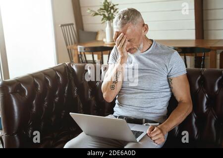 Frustrated middle aged man touching his head and feeling stressed while sitting with laptop on sofa in the living room and working remotely Stock Photo