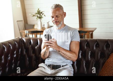 Texting message. Middle aged caucasian man in casual clothes using his smartphone while sitting on sofa in the living room Stock Photo