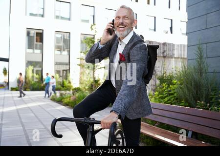 Happy worker. Cheerful middle aged businessman in stylish suit smiling while talking on the phone, standing with his bicycle outdoors Stock Photo