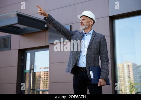 Improving the process. Confident middle aged architect, engineer manager wearing helmet pointing and looking away while standing outdoors and holding Stock Photo