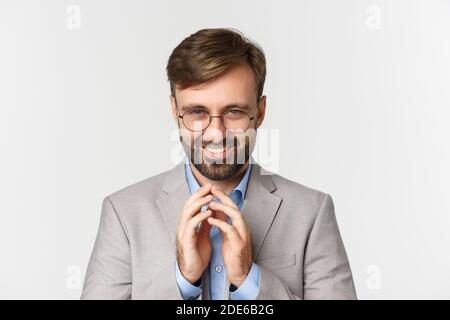 Close-up of bearded businessman in glasses and suit, scheming something, smiling devious and having perfect plan, standing over white background Stock Photo