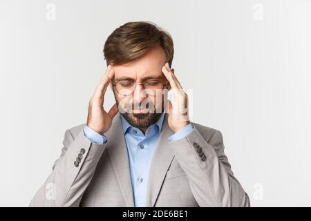Close-up of tired office worker in gray suit and glasses having severe headache, touching head and grimacing from pain in head, standing over white Stock Photo