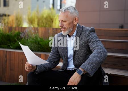Focused elegant middle aged businessman holding papers, reading financial statistics while sitting outdoors Stock Photo