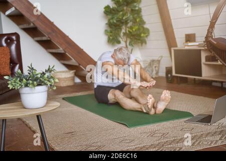 Sporty healthy middle aged man doing stretching exercises while sitting on yoga mat in the living room, watching online video tutorial on laptop Stock Photo