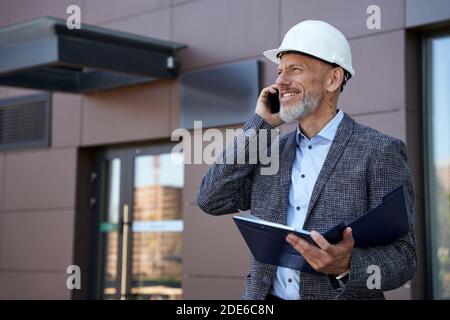 Discussing ideas. Cheerful mature architect, engineer manager wearing helmet looking away, talking on the phone while holding a blueprint, standing Stock Photo