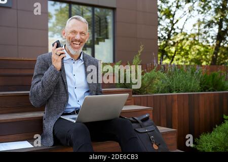 Improve your business. Joyful mature businessman smiling, making a call using his mobile phone while working on the laptop, sitting outdoors on the Stock Photo