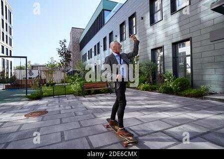 Life in motion. Full length shot of joyful stylish mature businessman smiling while taking a selfie using his mobile phone, riding his skateboard near Stock Photo