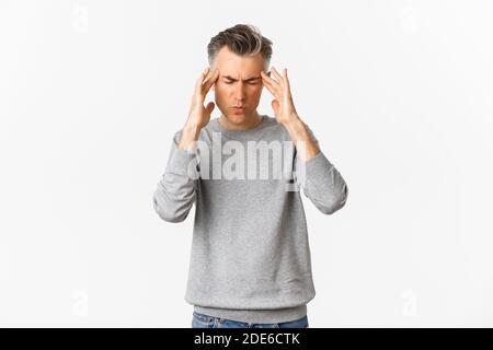 Portrait of middle-aged man in grey sweater having a headache, touching head and grimacing from painful migraine, standing over white background Stock Photo