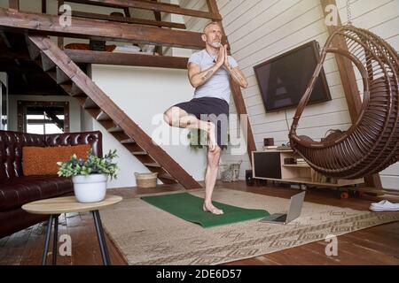 Healthy and calm middle aged man practicing yoga at home while watching online video tutorial on his laptop Stock Photo