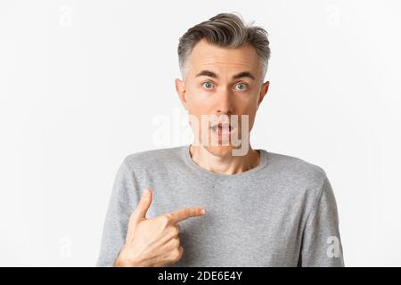 Close-up of confused middle-aged man, pointing at himself and gasping, standing over white background Stock Photo