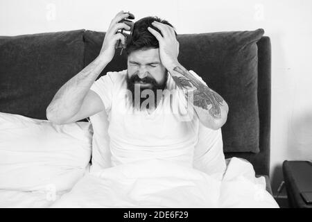 My clock is running late. Late riser suffer headache. Stressed hipster hold alarm clock in bed. Bearded man wake up late in morning. Keeping late hours. Wake time after sleep. Stock Photo