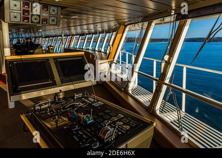 Two radar and navigation screens glow at the control desk on the bridge of the Pont Aven of Brittany Ferries. Through the window front, the green coastline is visible in the morning light as the ferry enters the port of Cork, Ireland; several crew members talk in the background. Stock Photo