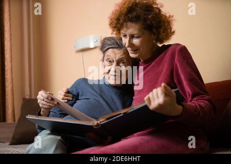 Positive aged ladies looking album photos sitting sofa at home, cheerful friends. Senior woman and her mature nurse watching photo album. Granny showi Stock Photo