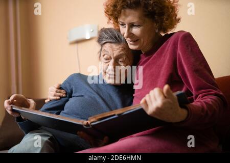 Positive aged ladies looking album photos sitting sofa at home, cheerful friends. Senior woman and her mature nurse watching photo album. Granny showi Stock Photo