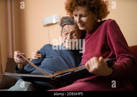 Positive aged ladies looking album photos sitting sofa at home, cheerful friends. Senior woman and her mature nurse watching photo album. Granny showi Stock Photo